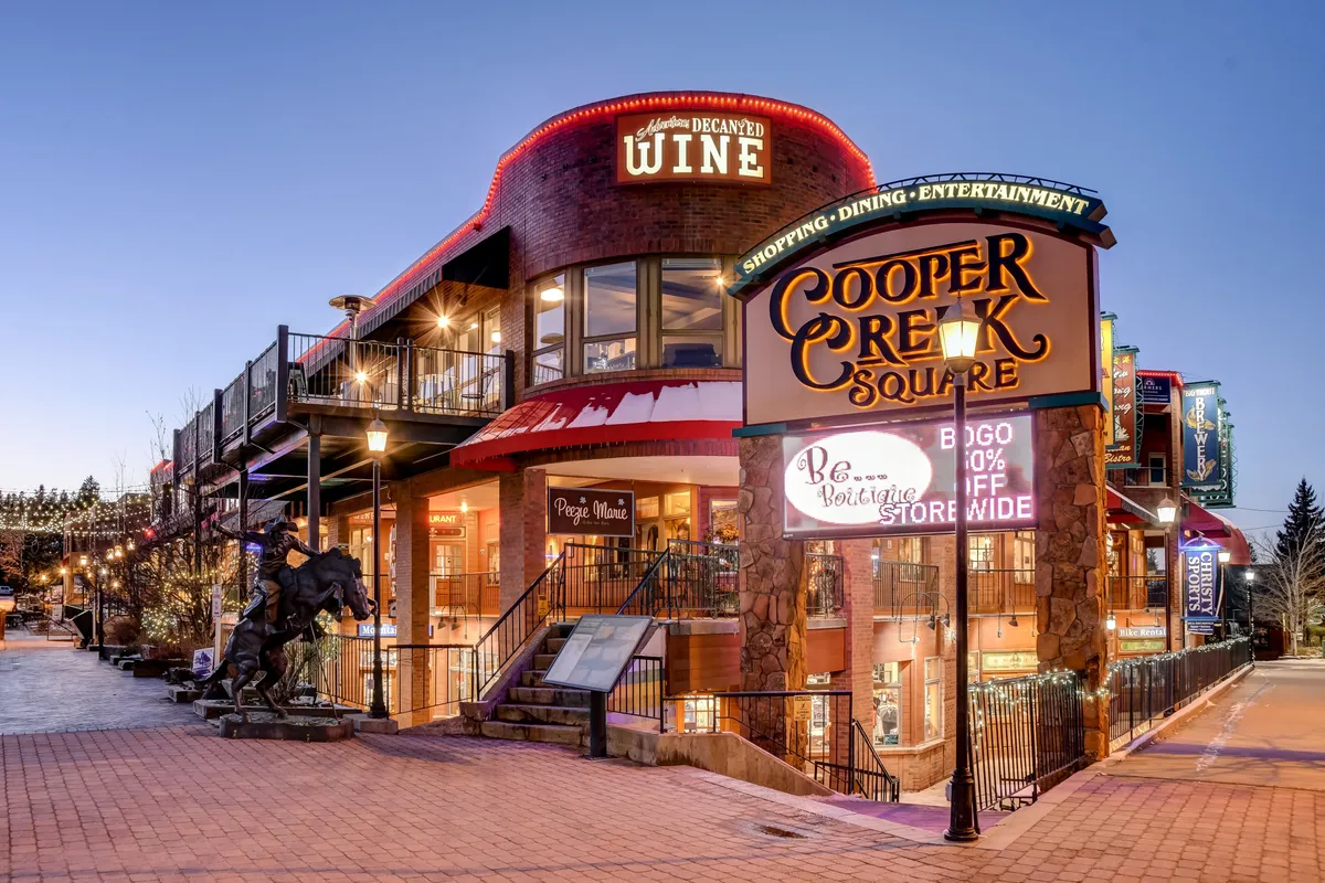 A lively outdoor plaza at Cooper Creek Square, featuring signage for shops and wine bars, an outdoor seating area, and a striking horse sculpture. The atmosphere is bustling, with people shopping, dining, and enjoying social moments amid the colorful, vibrant setting.