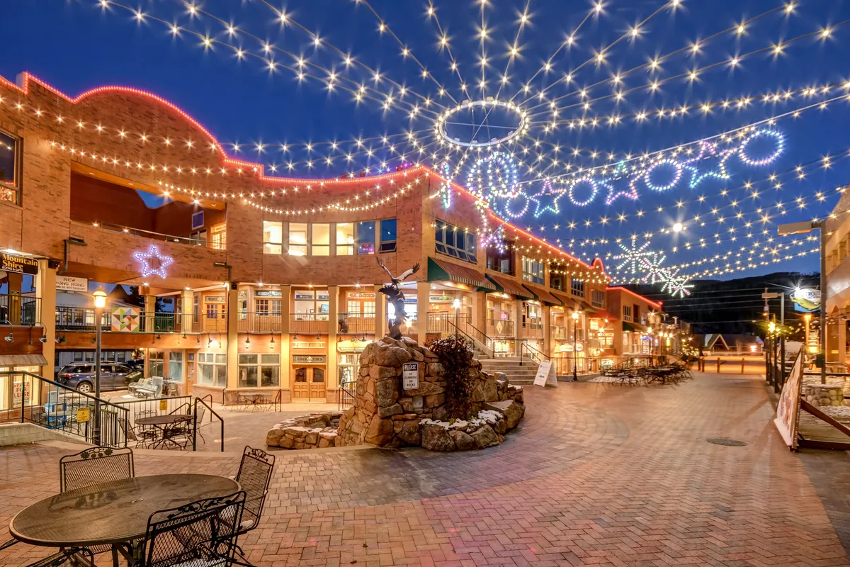 A cheerful exterior scene featuring inviting string lights overhead, a decorative stone fountain, and buildings with large windows. The outdoor seating areas suggest a vibrant community environment, perfect for gathering and enjoying local amenities.