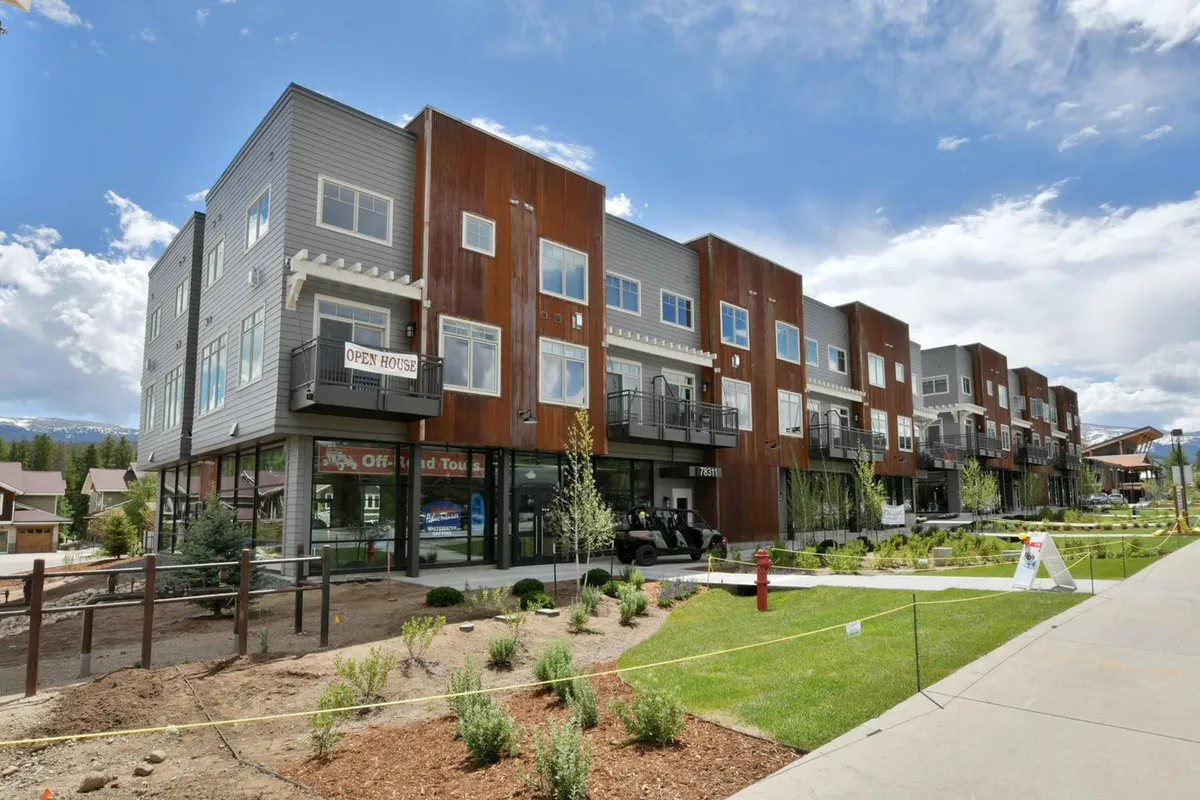 This exterior image highlights the modern architecture of the condo-style building surrounded by lush green landscaping. The inviting outdoor spaces and open house sign convey a welcoming community atmosphere and hint at the unique blend of private home comfort within a shared, resort-like environment.