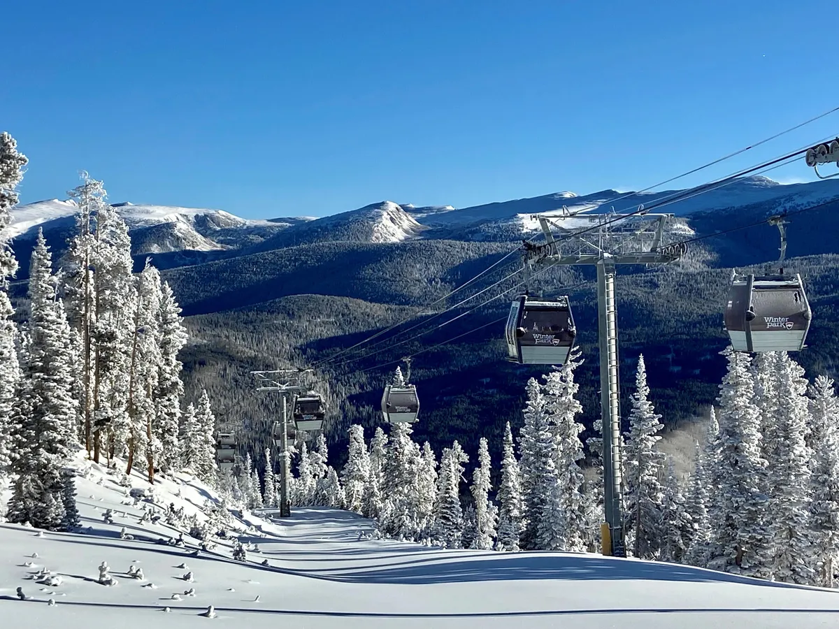 An exterior shot capturing ski lift gondolas ascending over snow-covered trees with a dramatic mountain range in the background. The scene evokes the excitement of winter sports and breathtaking alpine views.