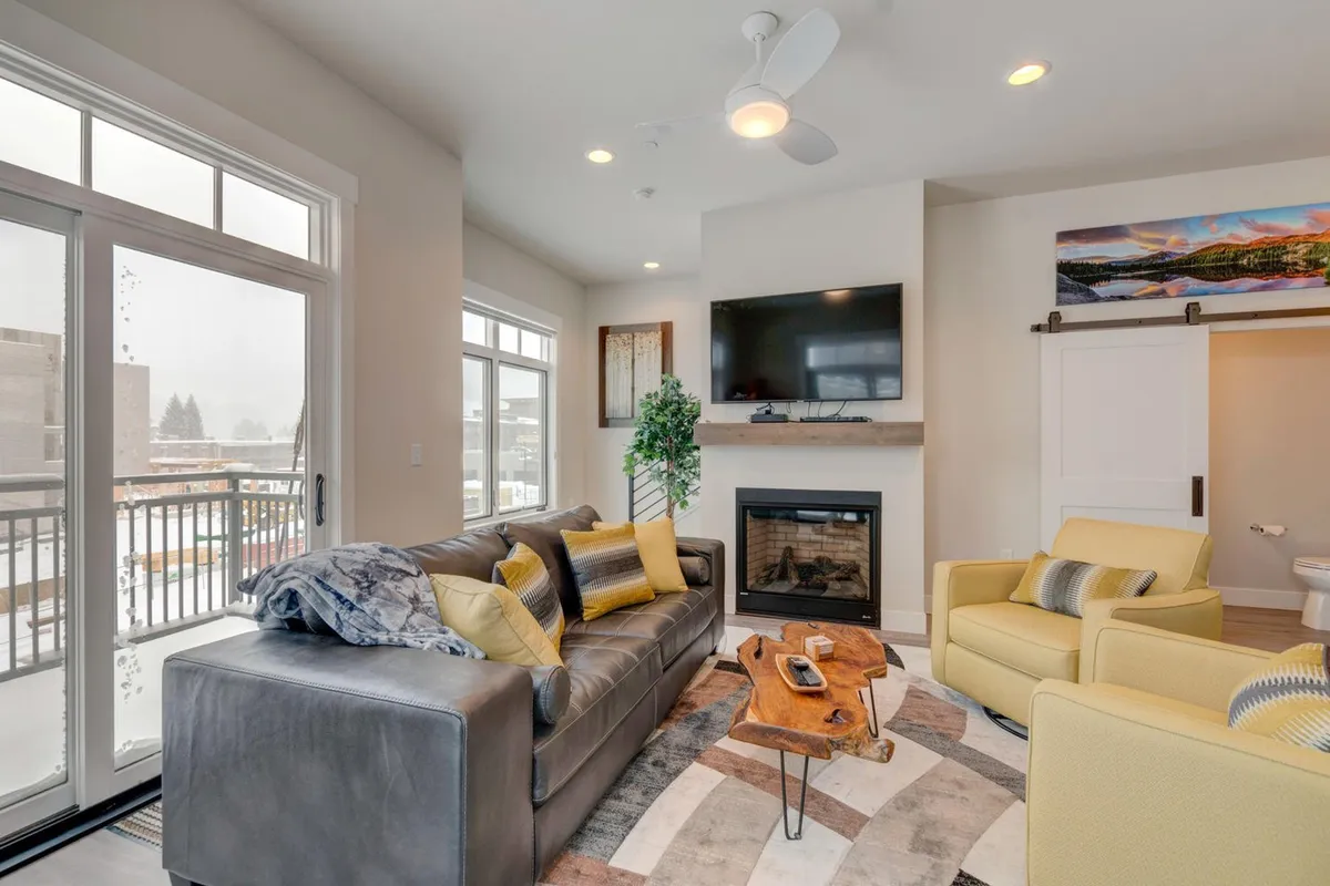 This image showcases a welcoming living room featuring a plush sofa facing a modern fireplace, a wall-mounted television, and vibrant yellow chairs. The space is bathed in natural light from large windows, with a coffee table at the center, creating an inviting spot for relaxation or entertainment.
