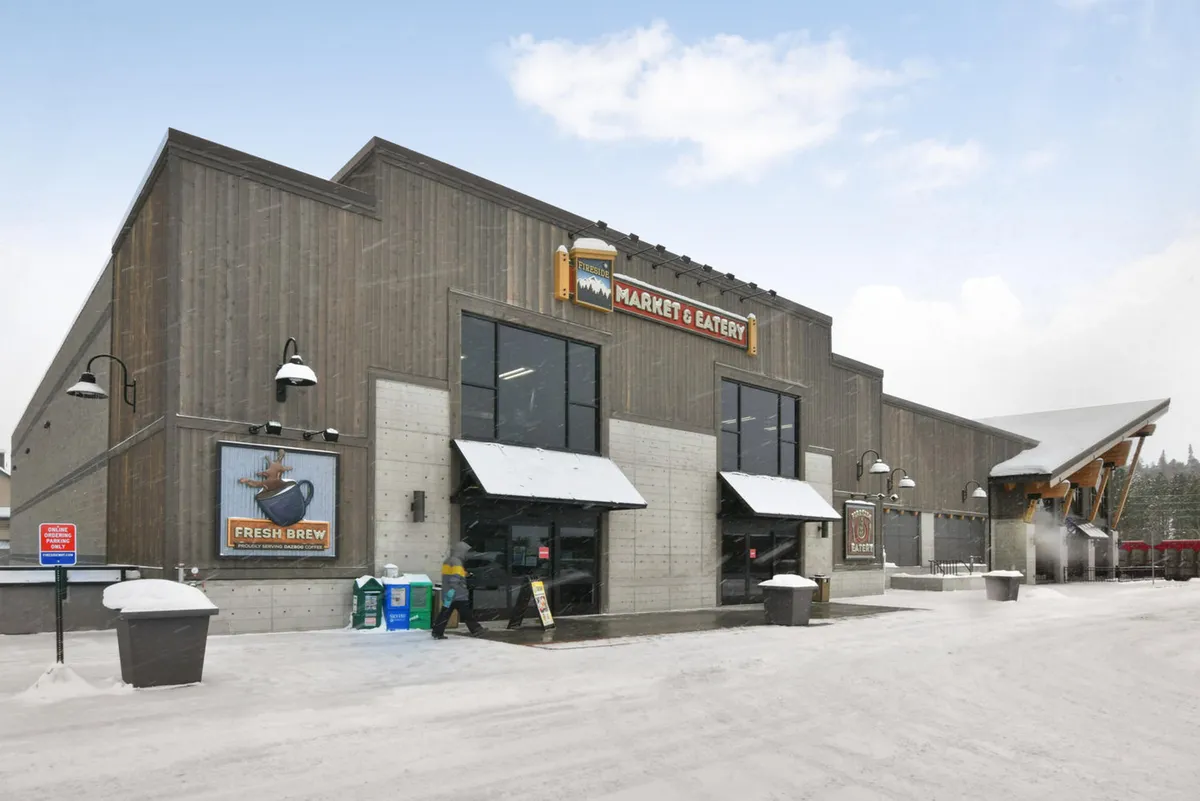 This exterior image features the main entrance of the Market & Eatery, with glass doors welcoming visitors, a prominent 'Market & Eatery' sign, and a snow-covered pathway leading up to the entrance. People are seen shopping for fresh produce, enjoying a coffee or meal, and interacting with one another, all set within a warm and inviting atmosphere despite the chilly weather.
