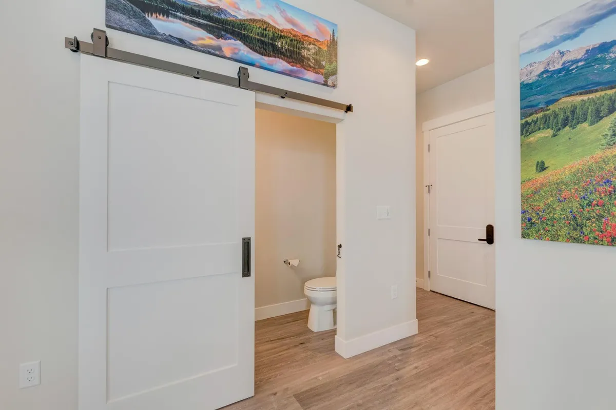 A welcoming upstairs hallway featuring a sliding barn door, decorative mountain landscape artwork, a toilet area, and colorful floral accents. The bright space hints at personal touches and easy access to bathroom facilities.