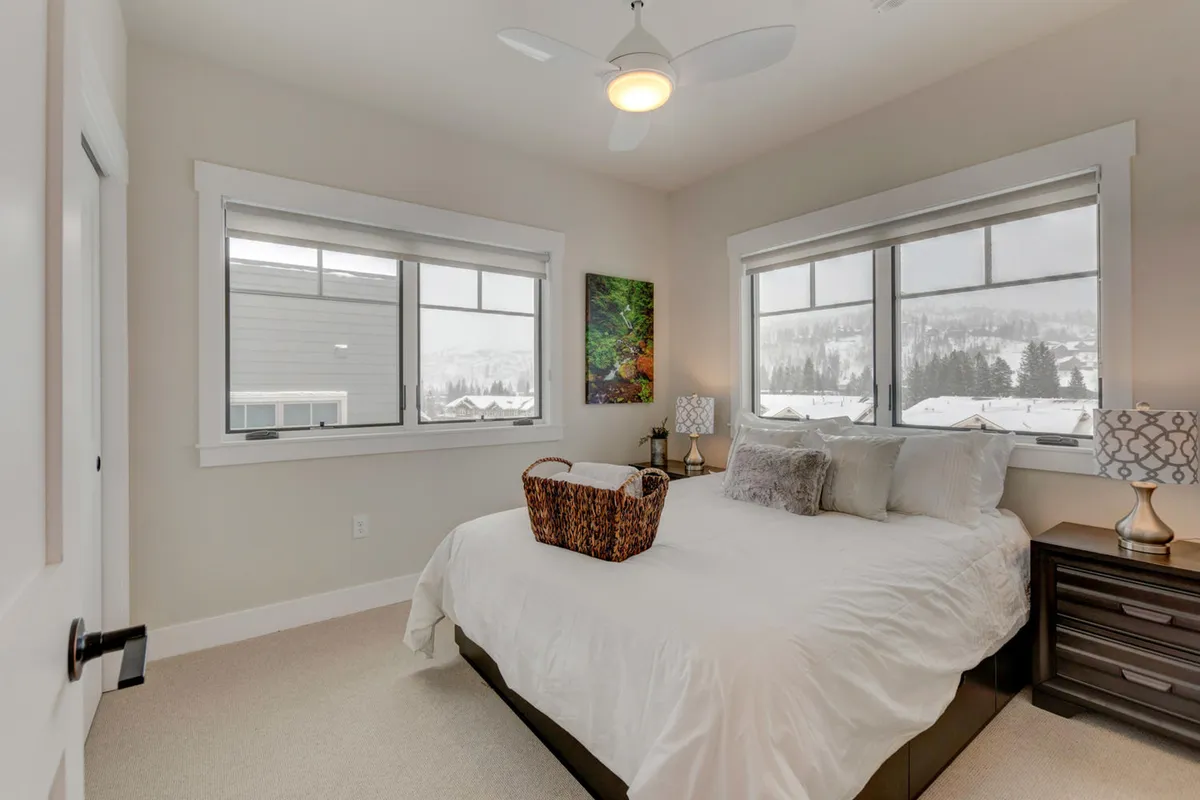 A serene bedroom with a comfortable bed, nightstand, lamp, and large windows letting in soft natural light. Artwork above the bed and a cozy atmosphere suggest a restful, private retreat ideal for relaxation after a busy day.