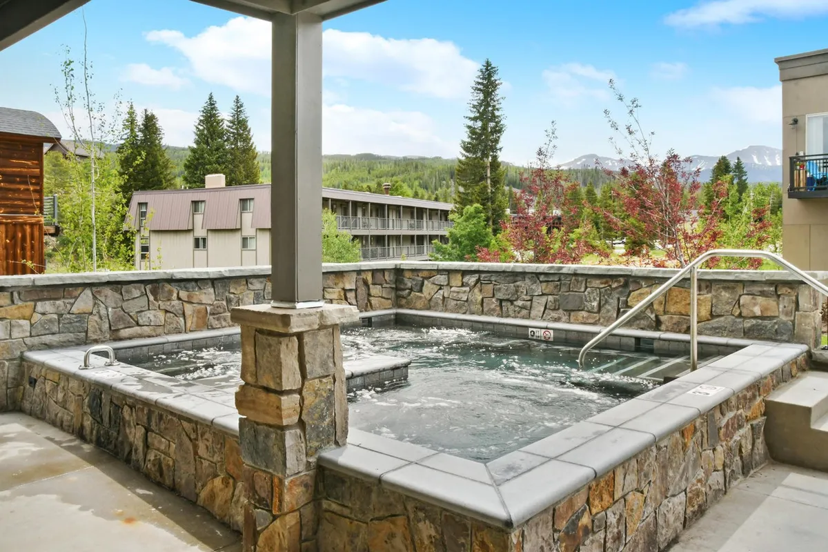 An inviting community hot tub set against a tranquil landscape, framed by stone walls and surrounded by natural scenery. The image captures people relaxing in the warm water and enjoying views of the outdoors, suggesting a social and rejuvenating atmosphere.