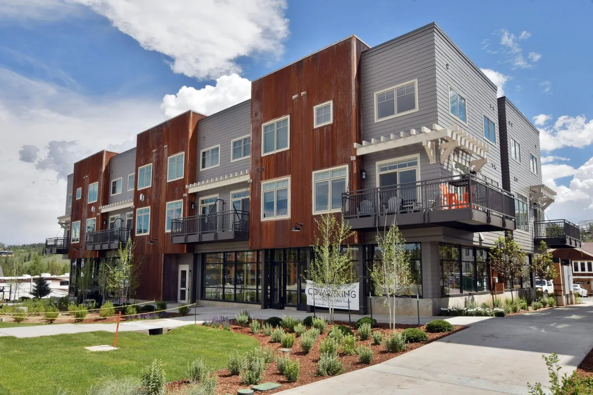 The exterior of a modern building with inviting balconies and a beautifully landscaped garden, where people are seen relaxing outdoors and socializing. The setting is warm and sunlit, conveying the pleasures of summer living and community gatherings.