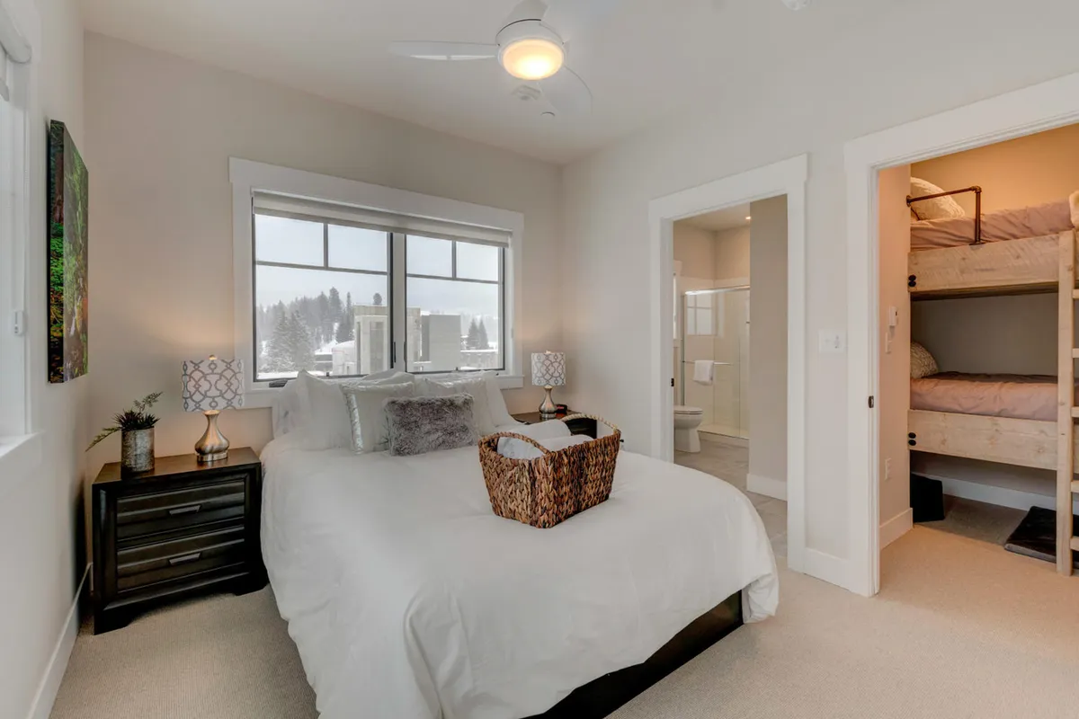 An inviting bedroom with a plush bed adorned with decorative pillows, bunk beds in the corner, soft lighting from lamps, and a window framing a snowy landscape. The room looks ideal for families or groups, offering both relaxation and space for children.